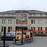 Zielony budynek dwupiętrowy na rynku, widoczny jest parking oraz wystawa przed budynkiem