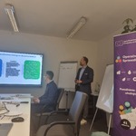 Na zajęciu widać dwie osoby, jedna prowadzi spotkanie i opowiada o tym co jest na wyświetlanym slajdzie, widać stół praz po prawej stronie baner