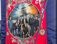 Zdjęcie sztandaru , koloru czerwonego , na pierwszym planie sześciu mężczyzn i koń, w tle szyb kopalni.