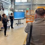 Hala produkcyjna, widoczne 3 osoby i ruchomy ekran z prezentacją.
