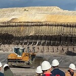Na pierwszym planie maszyna górnicza w tle pozostałości po kopalni odkrywkowej, hałdy, wykopaliska koloru piasku. U dołu zdjęcia ludzie w białych kaskach.