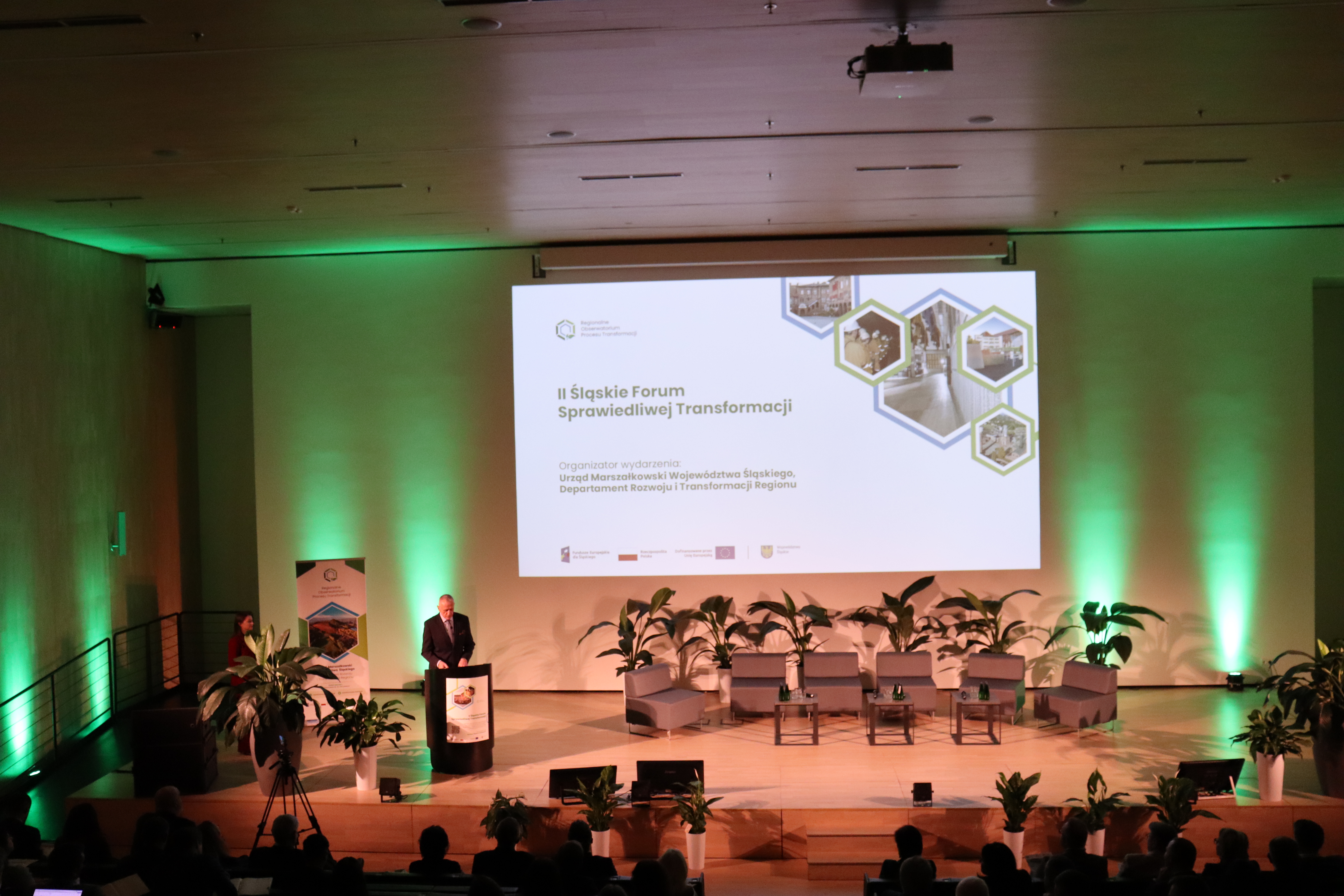 Na zdjęciu Marszałek Województwa Śląskiego Leszek Pietraszek otwiera II Śląskie Forum Sprawiedliwej Transformacji. Widoczny jest ekran ze slajdem informującym, że organizatorem II Śląskiego Forum Sprawiedliwej Transformacji jest Departament Rozwoju i Transformacji Regionu Urzędu Marszałkowskiego Województwa Śląskiego. Na scenie są fotele dla panelistów, małe stoliczki, w tle - baner Regionalnego Obserwatorium Procesu Transformacji oraz kwiaty doniczkowe.