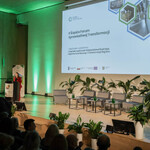 Na zdjęciu widoczna jest część widowni wraz z uczestnikami. Dodatkowo widać scenę. Przy mównicy osoba moderująca Forum. Widoczny jest ekran ze slajdem informującym, że organizatorem II Śląskiego Forum Sprawiedliwej Transformacji jest Departament Rozwoju i Transformacji Regionu Urzędu Marszałkowskiego Województwa Śląskiego. Na scenie są fotele dla panelistów, małe stoliczki, w tle - baner Regionalnego Obserwatorium Procesu Transformacji oraz kwiaty doniczkowe.