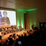 Na zdjęciu widoczna jest część widowni wraz z uczestnikami. Dodatkowo widać scenę i ekran, na którym wyświetlany jest materiał filmowy, w którym występuje Pani Emma Toledano Laredo, Dyrektor w Dyrekcji Generalnej ds. Polityki Regionalnej i Miejskiej Komisji Europejskiej. Na scenie widać fotele, małe stoliczki, kwiaty doniczkowe. Prowadząca Forum stoi na scenie, z boku.