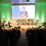 Na zdjęciu widoczna jest część widowni wraz z uczestnikami. Dodatkowo widać scenę i ekran, na którym wyświetlany jest materiał filmowy, w którym występuje Pani Emma Toledano Laredo, Dyrektor w Dyrekcji Generalnej ds. Polityki Regionalnej i Miejskiej Komisji Europejskiej. Na scenie widać fotele, małe stoliczki, kwiaty doniczkowe. Prowadząca Forum stoi na scenie, z boku. 
