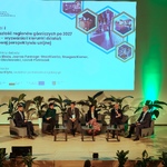 Na zdjęciu  widać scenę, na której jest pięciu prelegentów Panelu 1 i moderator siedzący na fotelach. Na ekranie jest slajd z tytułem panelu: „Panel I: Przyszłość regionów górniczych po 2027 roku – wyzwania i kierunki działań w nowej perspektywie unijnej” oraz imionami i nazwiskami prelegentów i moderatora. Na scenie są także małe stoliczki, kwiaty doniczkowe.
