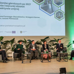 Na zdjęciu z bliska widać scenę, na której jest pięciu prelegentów Panelu 1 i moderator siedzący na fotelach. Na ekranie jest slajd z tytułem panelu: „Panel I: Przyszłość regionów górniczych po 2027 roku – wyzwania i kierunki działań w nowej perspektywie unijnej” oraz imionami i nazwiskami prelegentów i moderatora. Na scenie są także małe stoliczki, kwiaty doniczkowe. 