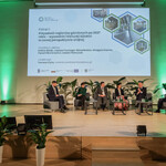 Na zdjęciu  widać scenę, na której jest pięciu prelegentów Panelu 1 i moderator siedzący na fotelach. Na ekranie jest slajd z tytułem panelu: „Panel I: Przyszłość regionów górniczych po 2027 roku – wyzwania i kierunki działań w nowej perspektywie unijnej” oraz imionami i nazwiskami prelegentów i moderatora. Na scenie są także małe stoliczki, kwiaty doniczkowe, a w tle - baner Regionalnego Obserwatorium Procesu Transformacji. 