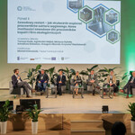 Na zdjęciu  widać scenę, na której jest sześciu prelegentów Panelu 2 i moderator siedzący na fotelach. Na ekranie jest slajd z tytułem panelu: „Panel II: Zawodowy restart – jak skutecznie wspierać pracowników sektora węglowego. Nowe możliwości zawodowe dla pracowników kopalń i firm okołogórniczych” oraz imionami i nazwiskami prelegentów i moderatora. Na scenie są także małe stoliczki, kwiaty doniczkowe. 