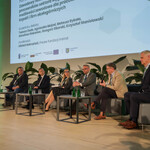 Na zdjęciu z bliska widać scenę, na której jest sześciu prelegentów Panelu 2 i moderator siedzący na fotelach. Dodatkowo część ekranu, na którym jest slajd z tytułem panelu: „Panel II: Zawodowy restart – jak skutecznie wspierać pracowników sektora węglowego. Nowe możliwości zawodowe dla pracowników kopalń i firm okołogórniczych” oraz imionami i nazwiskami prelegentów i moderatora. Na scenie są także małe stoliczki, kwiaty doniczkowe. 