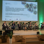 Na zdjęciu  widać scenę, na której jest sześciu prelegentów Panelu 2 i moderator siedzący na fotelach. Na ekranie jest slajd z tytułem panelu: „Panel II: Zawodowy restart – jak skutecznie wspierać pracowników sektora węglowego. Nowe możliwości zawodowe dla pracowników kopalń i firm okołogórniczych” oraz imionami i nazwiskami prelegentów i moderatora. Na scenie są także małe stoliczki, kwiaty doniczkowe. 