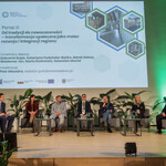 Na zdjęciu z bliska widać scenę, na której jest sześciu prelegentów Panelu 3 i moderator siedzący na fotelach. Na ekranie jest slajd z tytułem panelu: „Panel III: Od tradycji do nowoczesności – transformacja społeczna jako motor rozwoju i integracji regionu” oraz imionami i nazwiskami prelegentów i moderatora. Na scenie są także małe stoliczki, kwiaty doniczkowe. 