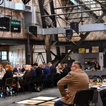 Spotkanie w przestrzeni poprzemysłowej: uczestnicy przy stołach, w tle metalowa konstrukcja i ceglane ściany