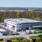 Panoramiczny widok zakładu produkcyjno-magazynowego firmy BOX HAUS z halą, częścią biurową, parkingiem, samochodami i składowanymi materiałami, otoczonego terenami zielonymi i zabudową przemysłową.