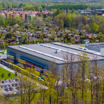 Widok z lotu ptaka na kompleks przemysłowy z dużą halą produkcyjno-magazynową, otoczony terenami zielonymi, ogródkami działkowymi i zabudową mieszkaniową.