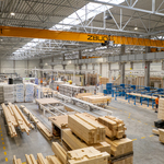 Wnętrze hali produkcyjnej firmy BOX HAUS z liniami technologicznymi do obróbki drewna, składowanymi elementami konstrukcyjnymi, wózkiem widłowym oraz suwnicą pomostową.