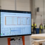 Panel sterowania maszyną produkcyjną z monitorem przedstawiającym projekt drewnianego elementu konstrukcyjnego; w tle wnętrze hali i pracownicy.
