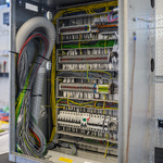 Otwarta metalowa szafa z gęsto rozmieszczonymi przewodami w różnych kolorach, listwami zaciskowymi, modułami elektrycznymi i rzędami zabezpieczeń. Po lewej stronie widoczna jest duża wiązka przewodów oraz elastyczny szary przewód, a całość znajduje się na stanowisku roboczym w hali.